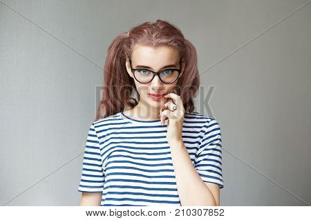 Studio shot of charming trendy looking pink haired European girl with blue eyes and two ponytails staring at camera in a playful manner holding finger at her lips posing at grey blank wall
