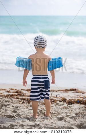Miami USA October 07 2017: Back view on cute toddler boy wearing safety inflatable armbands standing on the beach ready to jump into ocean or sea. Safety first. Vacations.