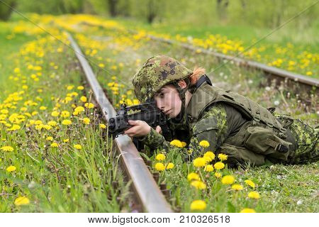 Portrait of armed woman with camouflage. Young female soldier observe with firearm on rail. Child soldier with gun in war green grass and yellow dandelions background. Military army people concept