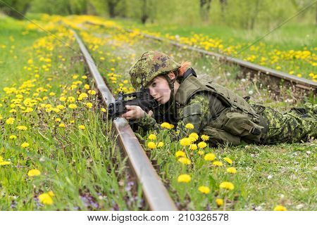 Portrait of armed woman with camouflage. Young female soldier observe with firearm on rail. Child soldier with gun in war green grass and yellow dandelions background. Military army people concept