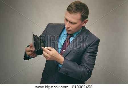 Young business man in a suit holds an empty wallet in his hand and looks for money in it. Pay the debt. Bankruptcy concept.
