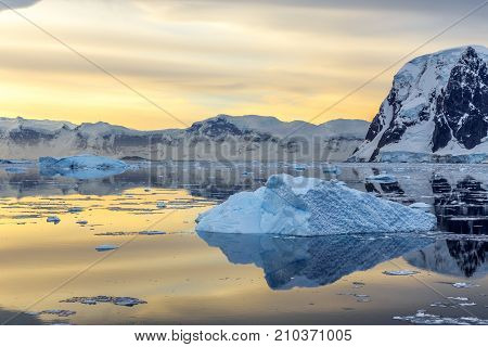 Cold Still Water Of Antarctic Lagoon With Drifting Blue Icebergs And Mountains In The Background, An