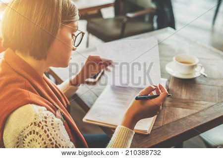 Side view. Young woman sits in cafe at a table, holding pen, reading documents. Online education, marketing. A student does homework. A businesswoman signs a contract. Startup, entrepreneur working.