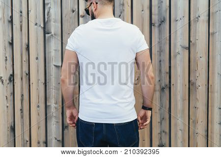 Summer day. Back view. Young bearded hipster man dressed in white t-shirt and sunglasses is stands outdoor against wood wall. Mock up. Space for logo, text, image.