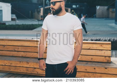 Summer day. Front view. Young bearded hipster man dressed in white t-shirt and sunglasses is stands on city street. Mock up. Space for logo, text, image. Instagram filter, film effect, bokeh effect.