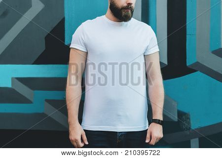 Summer day. Front view. Young bearded hipster man dressed in white t-shirt is stands outdoor against wall with graffiti. Mock up. Space for logo, text, image.