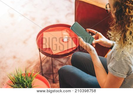 View from above. Young woman in glasses and striped T-shirt is sitting at home on couch and is using smartphone. Hipster girl is checking e-mail.