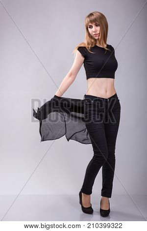 Beautiful young casual girl taking off black shirt and black pants wearing black top. Full body length standing portrait over gray background.