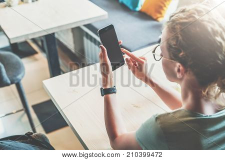 Rear view. Hipster girl is sitting in cafe at table, using digital gadget, looking on screen of smartphone, checking email. Businesswoman working online.