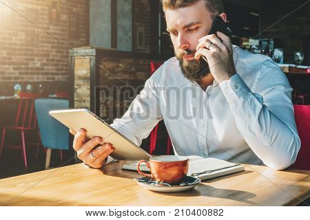 Young bearded businessman sits in cafe at table, talking on mobile phone, holding tablet computer.Man is working
