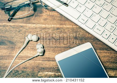 White Mobile Next To In-ear Headphones, Glasses And White Keyboard On Wooden Table