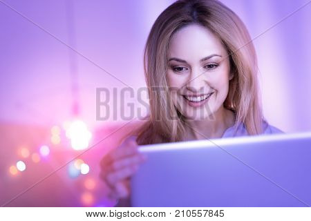 Nice talk. Lovely vigorous blond woman sitting with her laptop and smiling while talking to someone on the laptop
