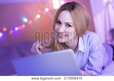Full of joy. Attractive happy young woman smiling and lying while working on the laptop
