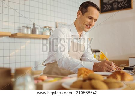 Successful entrepreneur. Smart nice handsome man standing in the kitchen and taking notes while working in his cafe