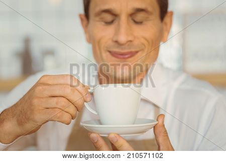 Cup of coffee. Selective focus of a cup with delicious drink being in hands of a cheerful delighted nice man while smelling it
