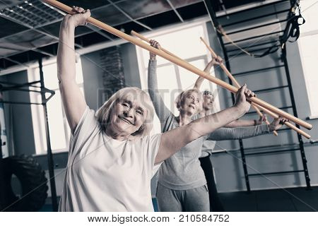 Sport brings joy. Three pleasant lively senior women standing one behind another in a gym and doing stretching exercises with wooden sticks while smiling happily