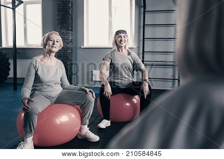 Being attentive. Two pleasant senior women sitting on stability balls with their legs spread and listening attentively to their personal trainer explaining them the next exercise