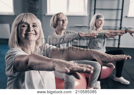 Together funnier. Three cheerful elderly women sitting on exercise balls, stretching out hands and smiling while one of them looking at the camera