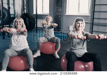 Healthy lifestyle. Three cheerful senior ladies sitting on exercise balls and stretching out hands with dumbbells while smiling at the camera