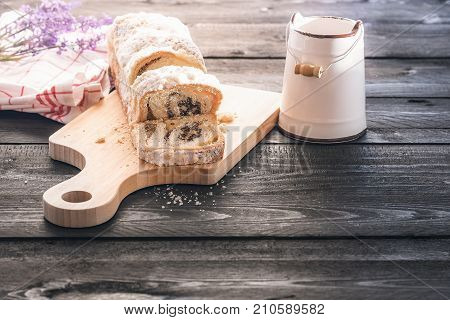 Delicious homemade sponge cake with poppy seed and walnuts sliced on a trencher near an antique bucket with milk on a black wooden table in the morning light.