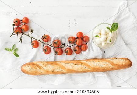 Baguette with banch of cherry-tomatoes, basil and bowl of mozzarella cheese on rustic white wooden backdrop, top view, copy space
