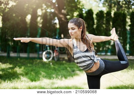 Close up of young female doing dandayamana dhanurasana in a city park relaxing striving for a perfect body. Outdoor yoga concept.
