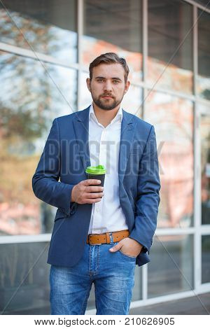 Attractive business man with beard drinking coffee and posing on the camera on the street. Panoramic wondows on the background. Close-up of man.