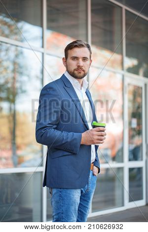 Attractive business man with beard drinking coffee and posing on the camera on the street. Panoramic wondows on the background. Close-up of man.
