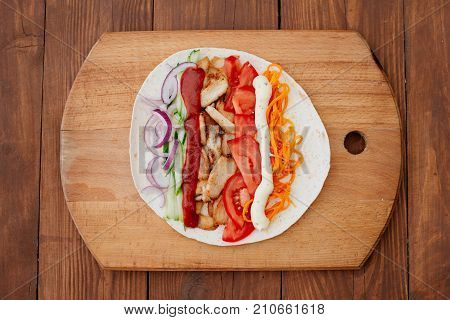 The process of preparing chiken shawarma. Ingredients sliced lie on the kitchen board. Fast food. Eastern food. Dishes of oriental cuisine lying on pita bread and decorated with greens and vegetables.