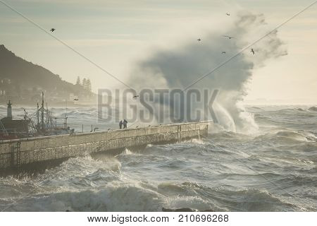 People risk their lives at Kalk Bay as a huge winter storm ocean wave crashes over the harbor wall.