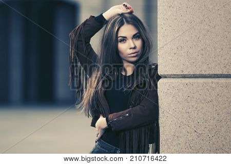 Happy young woman standing at the wall in city street. Stylish fashion model in leather fringe suede jacket 
