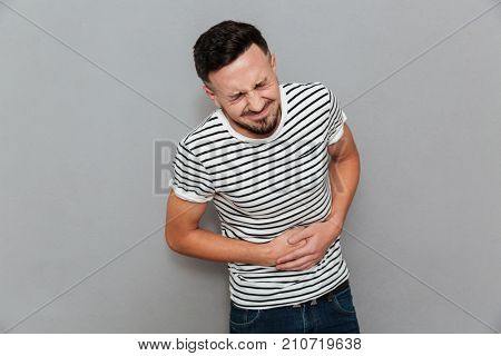 Image of displeased young man with painful feelings holding his belly isolated over grey background. Eyes closed.