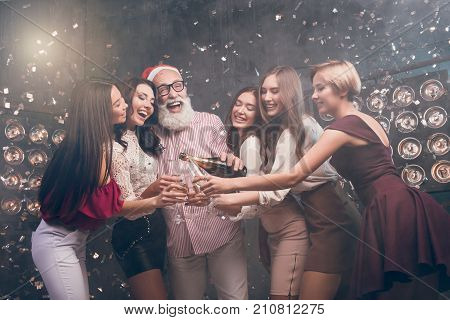 The bearded man in Santa's hat pours champagne into girl's glasses and smile. Modern Santa in Christmas hat have a fun with girls in the club with confetti. Happy New Year 2018