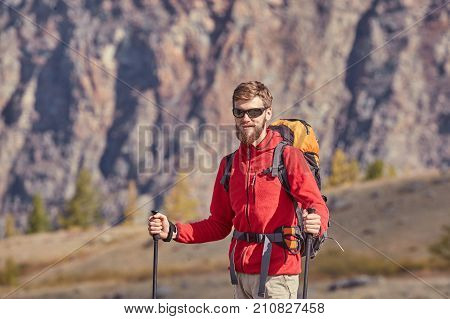 Tall backpacker with beard and sunglasses hold poles in hand. Sunny spring daybreak in rocky mountains. Hiker with backpack stand on rocky view point above valley.