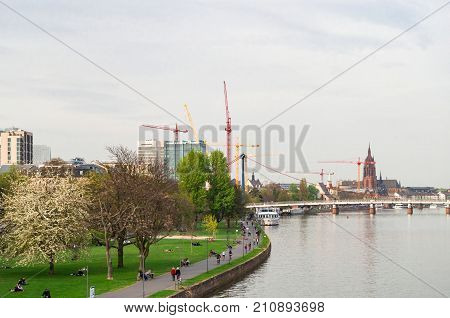 View of the Main promenade in Frankfurt with walking and resting people and the Holbeinsteg- bridge, the Imperial Cathedral of Saint Bartholomew (Kaiserdom) and some building cranes in the background. Frankfurt (Main), Germany - April 1 2014.