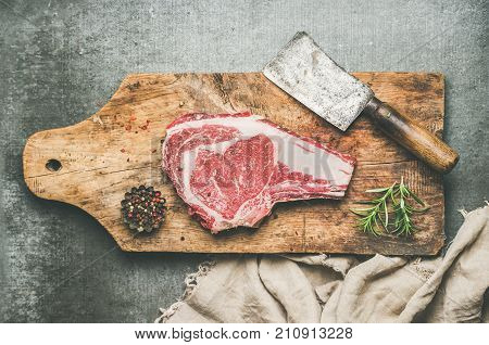Flat-lay of raw prime beef meat dry-aged steak rib-eye on bone with seasoning and chopper knife on wooden cutting board over grey concrete background, top view. Meat high-protein dinner concept
