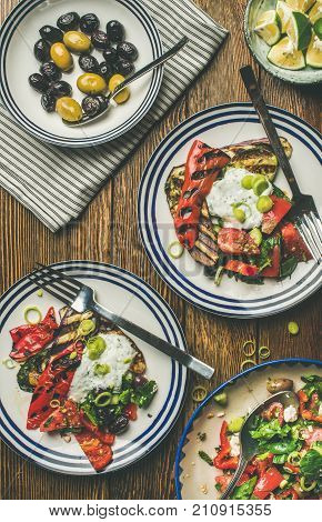 Flat-lay of healthy dinner table setting. Green salad, grilled vegetables with yogurt sauce, pickled olives on plates, lemon water over wooden background, top view. Clean eating, vegan food concept