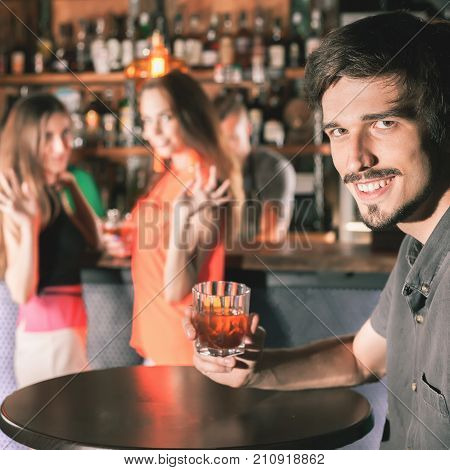 Handsome drunk man sitting at bar, drinking cocktail and looking at two attractive girls. He wants to get to know them, but does not know how. Nightclub. Date