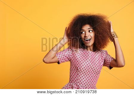 Close-up portrait of charming african woman in dress touching her afro hairstyle, looking aside, isolated on yellow background