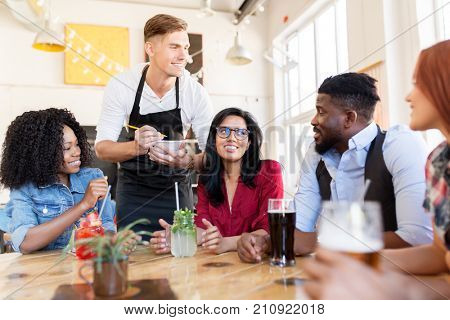 service and people concept - group of happy international friends with drinks and waiter with notepad receiving order at bar or restaurant