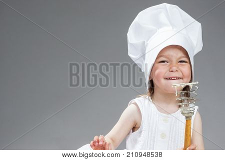 Funny Cooking Concepts. Portrait of Smiling Little Caucasian Girl in Cook Uniform Posing with Whisk Covered with Flour. Against Gray. Horizontal Image