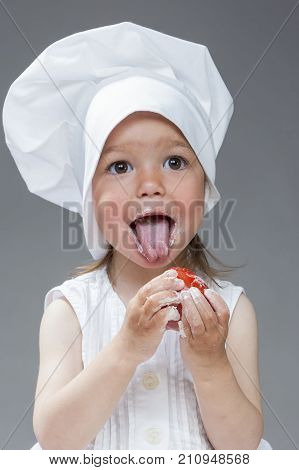 Cooking Ideas. Portrait of Funny Caucasian Little Girl in Cook Uniform Posing Fresh Tomato and with Smudgy Face Against Gray Background. Vertical Image Orientation