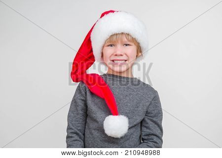 Little blond emotional boy in a festive red hat in a gray jacket. Little Santa Claus on a gray background.