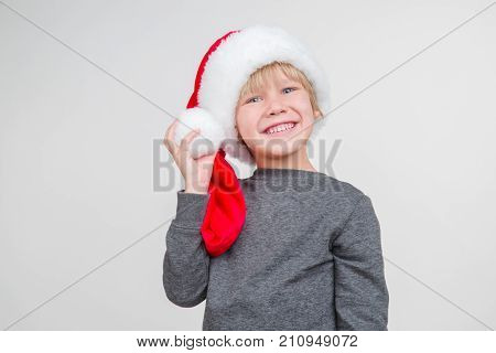 Little blond emotional boy in a festive red hat in a gray jacket. Little Santa Claus on a gray background.