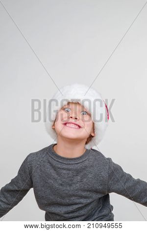 Little blond emotional boy in a festive red hat in a gray jacket. Little Santa Claus.