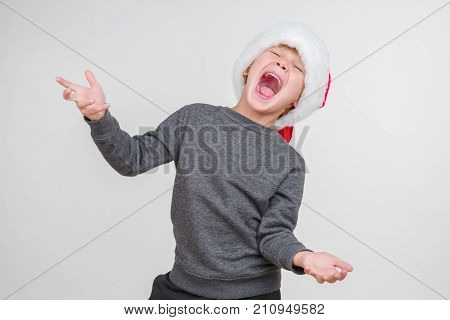 Little blond emotional boy in a festive red hat in a gray jacket. Little Santa Claus.