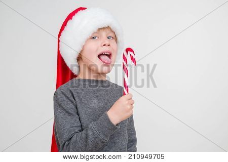 Little blond emotional boy in a festive red hat in a gray sweatshirt with a sweet stick. Little Santa Claus with lollipop on a gray background.