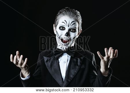 Portrait of a child boy with carnival make-up of a skeleton wearing elegant tail-coat. Studio shot over black background. Halloween. Dia de los muertos. The Day of the Dead.