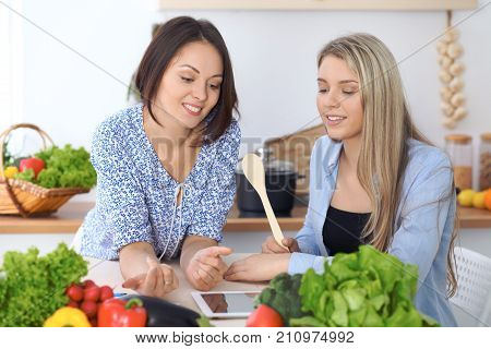 Two young happy women making online shopping for making menu by tablet computer. Friends cooking in the kitchen.