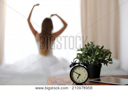 Alarm clock standing on bedside table has already rung early morning to wake up woman is stretching in bed in background. Early awakening, not getting enough sleep, oversleep concept.